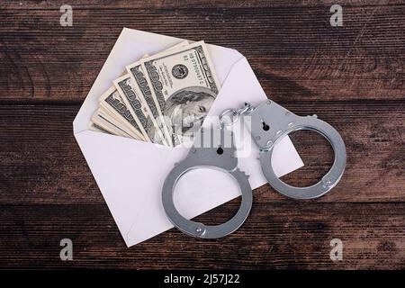 Handschellen, die auf einem offenen weißen Umschlag mit Dollars liegen, Nahaufnahme auf dunklem Hintergrund Stockfoto