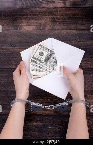 Hände mit Handschellen halten einen offenen weißen Umschlag mit Dollars auf einem dunklen Holzhintergrund Stockfoto