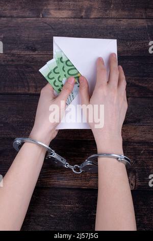 Hände mit Handschellen halten einen offenen Umschlag mit Euroscheinen auf einem dunklen Holzhintergrund Stockfoto