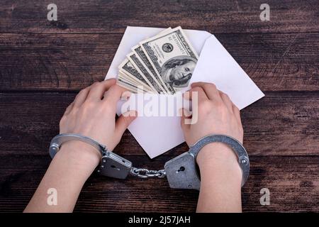 Die Hände mit Handschellen liegen auf einem offenen weißen Umschlag mit Dollars auf einem dunklen Holzhintergrund Stockfoto