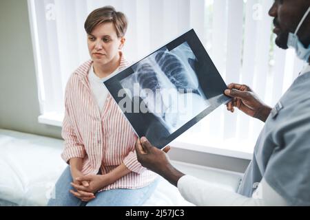 Schwarzer Arzt analysiert Röntgenbild einer kovidierten Patientin in der Lunge, während er mit einer Patientin über die Behandlung spricht Stockfoto
