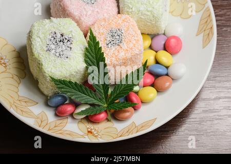 Süßes Marihuana-Dessert. Türkische Delight und bunte Süßigkeiten Konfetti mit grünen Cannabisblatt Nahaufnahme. Layout von Unkrautfutter mit CBD-Öl auf weißem Platt Stockfoto