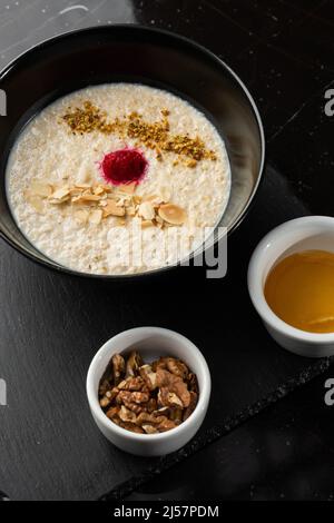 Grießbrei zum Frühstück Hafer mit Nüssen in schwarzer Schüssel serviert mit Honig isoliert auf schwarzem Stein Hintergrund. Hausgemachte Speisen. Leckeres Frühstück. Stockfoto