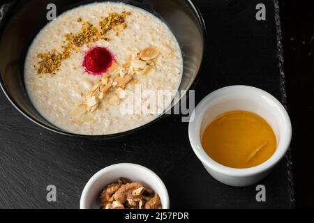 Grießbrei zum Frühstück Hafer mit Nüssen in schwarzer Schüssel serviert mit Honig isoliert auf schwarzem Stein Hintergrund. Hausgemachte Speisen. Leckeres Frühstück. Stockfoto