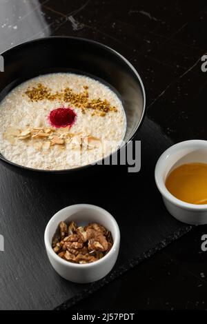 Grießbrei zum Frühstück Hafer mit Nüssen in schwarzer Schüssel serviert mit Honig isoliert auf schwarzem Stein Hintergrund. Hausgemachte Speisen. Leckeres Frühstück. Stockfoto