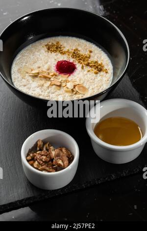 Grießbrei zum Frühstück Hafer mit Nüssen in schwarzer Schüssel serviert mit Honig isoliert auf schwarzem Stein Hintergrund. Hausgemachte Speisen. Leckeres Frühstück. Stockfoto