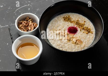 Grießbrei zum Frühstück Hafer mit Nüssen in schwarzer Schüssel serviert mit Honig isoliert auf schwarzem Stein Hintergrund. Hausgemachte Speisen. Leckeres Frühstück. Stockfoto