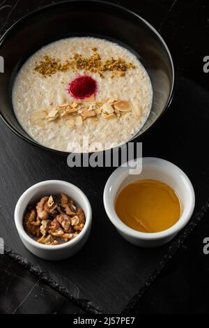 Grießbrei zum Frühstück Hafer mit Nüssen in schwarzer Schüssel serviert mit Honig isoliert auf schwarzem Stein Hintergrund. Hausgemachte Speisen. Leckeres Frühstück. Stockfoto