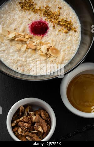 Grießbrei zum Frühstück Hafer mit Nüssen in schwarzer Schüssel serviert mit Honig isoliert auf schwarzem Stein Hintergrund. Hausgemachte Speisen. Leckeres Frühstück. Stockfoto