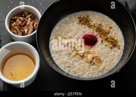 Grießbrei zum Frühstück Hafer mit Nüssen in schwarzer Schüssel serviert mit Honig isoliert auf schwarzem Stein Hintergrund. Hausgemachte Speisen. Leckeres Frühstück. Stockfoto