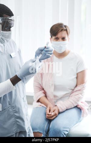 Konzentrierte schwarze Krankenschwester in schützendem Gesichtsschutz und Handschuhen, die eine Spritze vorbereiten, bevor sie gegen Coronavirus impft Stockfoto