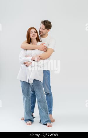 Lächelnder Mann in Jeans umarmt positive Freundin im Hemd auf weißem Hintergrund Stockfoto