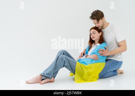 Junger Mann umarmt barfuß Freundin in ukrainischer Flagge auf weißem Hintergrund Stockfoto