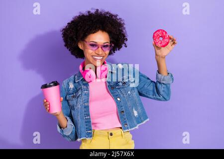 Foto von verträumt lustig junge Frau tragen Jeans Jacke Kopfhörer trinken Tee essen Kekse isoliert violette Farbe Hintergrund Stockfoto