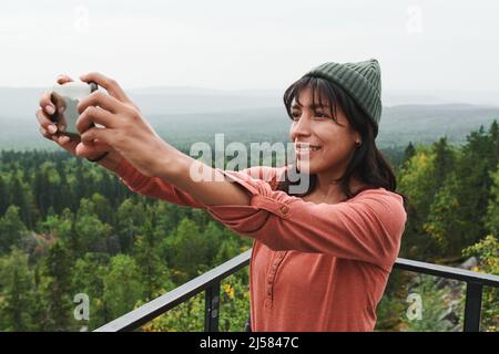 Lächelnde, hübsche junge Frau mit gemischtem Rennen im Hut, die auf der Brücke steht und sich auf dem Smartphone im Freien fotografiert Stockfoto