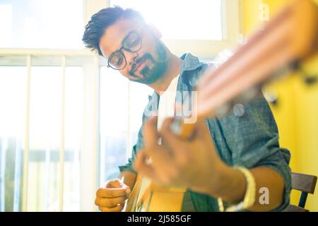 Fröhlicher, hübscher junger Mann, der Gitarre spielt und lächelt in einer neuen, gemütlichen Wohnung Stockfoto