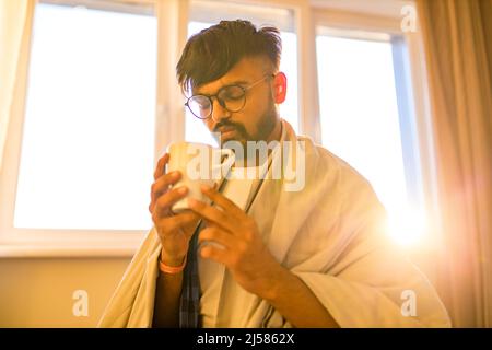 arabischer indischer Mann trinkt heißen Zitrone Dinder Masala Tee im Schlafzimmer am Morgen, während das Gefühl kalt mit einer Decke bedeckt Stockfoto