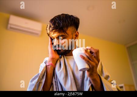 arabischer indischer Mann trinkt heißen Zitrone Dinder Masala Tee im Schlafzimmer am Morgen, während das Gefühl kalt mit einer Decke bedeckt Stockfoto