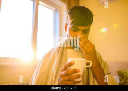 arabischer indischer Mann trinkt heißen Zitrone Dinder Masala Tee im Schlafzimmer am Morgen, während das Gefühl kalt mit einer Decke bedeckt Stockfoto