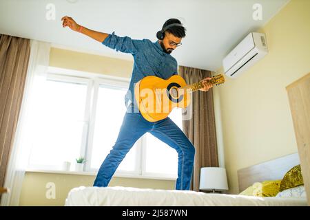 Fröhlicher, hübscher junger Mann, der Gitarre spielt und lächelt in einer neuen, gemütlichen Wohnung Stockfoto