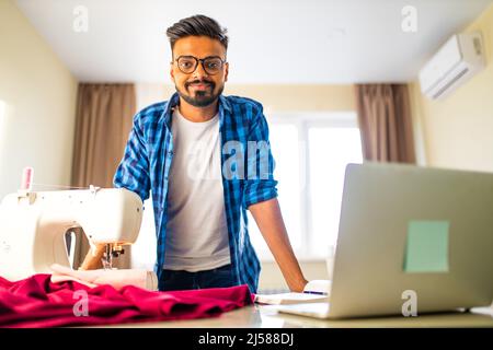 indische Schneider asiatische Mann Designer nähen Kleidung auf Nähmaschine neben Laptop Wohnung Arbeitsplatz Stockfoto