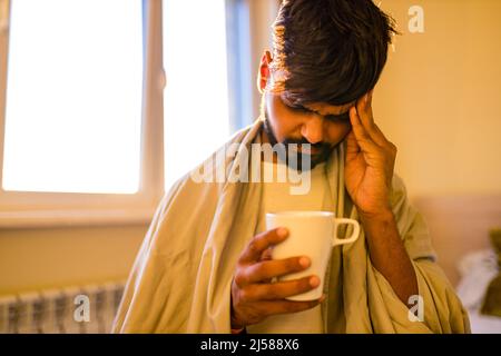 arabischer indischer Mann trinkt heißen Zitrone Dinder Masala Tee im Schlafzimmer am Morgen, während das Gefühl kalt mit einer Decke bedeckt Stockfoto