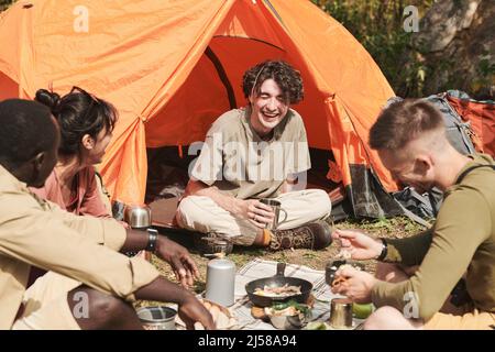 Gruppe fröhlicher junger multiethnischer Freunde von Wanderern, die beim Zelten lachen und zusammen essen Stockfoto