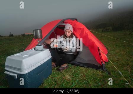 Schöne Lateinerin, die allein vor ihrem roten Zelt inmitten eines grünen Feldes sitzt und Tee aus ihrer Thermoskanne neben einem Kühler in einer bewölkten Sonne trinkt Stockfoto