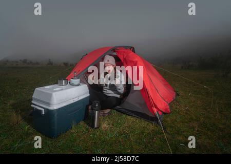 Schöne Lateinerin, die allein vor ihrem roten Zelt inmitten eines grünen Feldes sitzt und Tee aus ihrer Thermoskanne neben einem Kühler in einer bewölkten Sonne trinkt Stockfoto