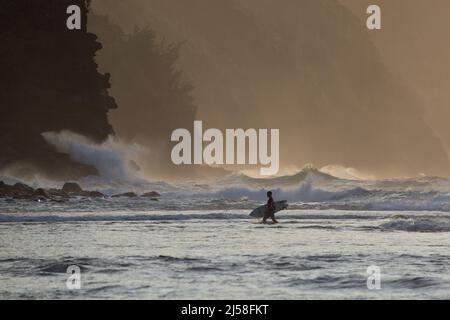 Am Ende des Tages kommt ein Surfer zum Ke'e Beach, während die Wellen heftig gegen die felsigen Klippen der Na Pali-Küste auf Kauai, Hawaii, krachen. Stockfoto