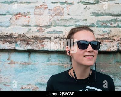 Porträt eines jungen attraktiven Mädchen in schwarzem T-Shirt und Sonnenbrille im Rock-Stil auf städtischen Hintergrund Musik mit Kopfhörern hören. Hipster in wir Stockfoto