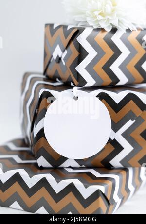 Geschenke mit leerem runden Geschenkanhänger aus nächster Nähe. Geschenkschachteln in geometrischem Chevron-Papier verpackt. Weihnachten, Neujahr, Geburtstag, Anniversary Label Mockup Stockfoto