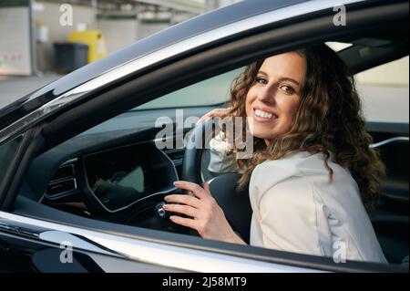 Porträt einer Geschäftsfrau mit einem Lächeln im Gesicht, die ihr Auto liebt. Lockiges, fröhliches Mädchen, das in der Kabine ihres Autos sitzt und die Hände hinter dem Lenkrad hält. Stockfoto