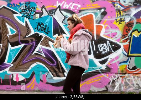 Person Frau, die an einem Wandgemälde auf dem Horten in der Brick Lane vorbei geht Shoreditch, der Bagel isst April 2022 London England Großbritannien KATHY DEWITT Stockfoto