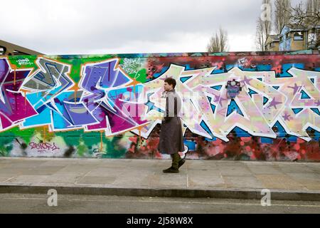 Mann Person trägt Trenchcoat Spaziergang durch bunte Malerei Wandbild auf horten Brick Lane Shoreditch Frühjahr 2022 London England UK KATHY DEWITT Stockfoto