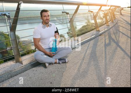 Ein gutaussehender, muskulöser Mann aus Europa, ein entschlossener Sportler, ein Sportler mit Kopfhörern, der sich nach dem Training im Freien in der Morgendämmerung entspannt, die Wasserflasche hält und sitzt Stockfoto