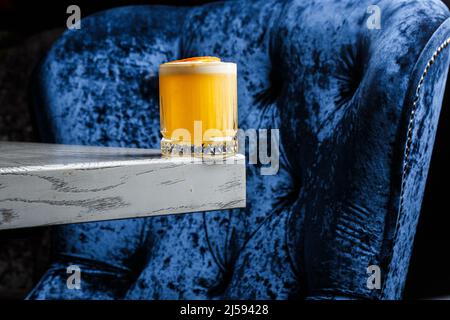 Ein Glas eines gelben Cocktails mit einem großen Eiswürfel aus weißem Schaumstoff, der mit orangefarbenen Chips auf dem blau verschwommenen Hintergrund verziert ist. Stockfoto
