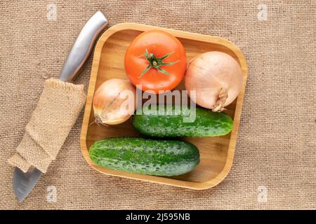 Eine Bio-Tomate, zwei Zwiebeln und zwei Gurken mit Bambusplatte und Messer auf Jute-Stoff, Makro, Draufsicht. Stockfoto