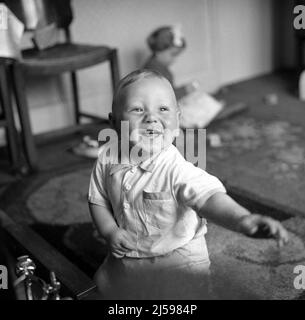 1962, historische Nahaufnahme eines glücklichen jungen Jungen, der in einem Vorderzimmer mit Blick auf den Markt spielt, England, Großbritannien. Ältere Schwester hinter ihm sitzt in der Ecke, Stockfoto