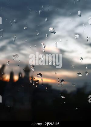 Es gibt viele Wassertropfen auf dem Glas. Verschwommene Gebäude und Abendhimmel hinter dem Glas Stockfoto