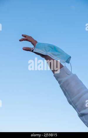Eine Hand, die im Hintergrund eine Maske gegen den Himmel hebt, nachdem sie die Pflicht zum Tragen abgeschlossen hat. Stockfoto