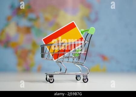 Spanische Nationalflagge in Miniatur-Einkaufswagen. Online-Shopping oder Versand, Duty-Konzept. Weltkarte im Hintergrund. Stockfoto