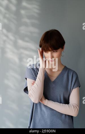 Verärgert junge kaukasische Frau mit kurzen Haaren massierenden Tempel, während sie unter Kopfschmerzen gegen graue Wand leiden Stockfoto