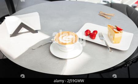 Klassischer Käsekuchen mit frischer Erdbeere mit einer Tasse Kaffee auf einem weißen Tisch. Nahaufnahme. Leckeres Frühstück. Stück Kuchen auf Teller, weiße Tasse auf Weiß Stockfoto