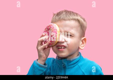 Ein süßer, lustiger Junge in einem blauen Pullover hält einen leuchtend rosa Donut in der Nähe seines Auges und schielt auf einem rosa Hintergrund. Fröhliches Kindheitskonzept. Süßes Essen für Stockfoto