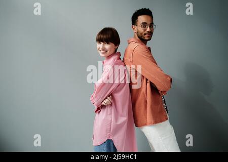 Porträt eines positiven interracial Paar von Modellen, die Rücken an Rücken stehen und die Arme auf der Brust gegen die graue Wand kreuzen Stockfoto
