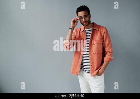 Portrait of content stilvoller junger afroamerikanischer Mann im Korallenshirt, der die Hand in der Jeans-Tasche hält und eine Einstellbrille trägt Stockfoto
