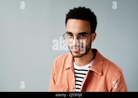 Porträt eines lächelnden, hübschen jungen Afro-Amerikaners in einer runden Brille und einem lässigen Hemd, das vor isoliertem Hintergrund steht Stockfoto