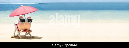 Rosa Sparschwein mit Sonnenbrille und Sonnenschirm auf Liegestuhl auf Sand am Strand Stockfoto