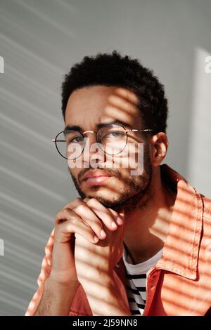 Porträt eines jungen schwarzen Mannes, der in runden Brillen Stirnrunzeln anstellt und im Schatten der Jalousien posiert Stockfoto
